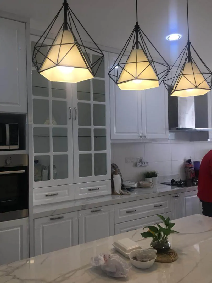 Dapur gaya shaker putih dengan kabinet pintu kaca, table top corak marmar dan tiga lampu gantung dawai hitam