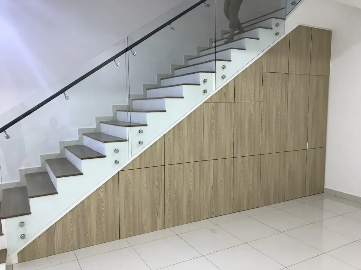 Under-stair cabinets in light oak wood-grain laminate following the slope of a staircase with glass railing